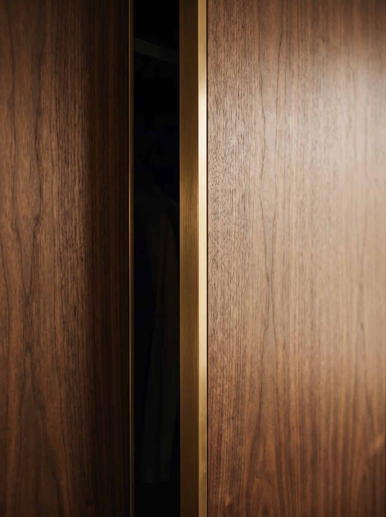 Close-up of floor-to-ceiling walnut wardrobe panels with a slim brass reveal strip and a garment glimpsed in the open gap.
