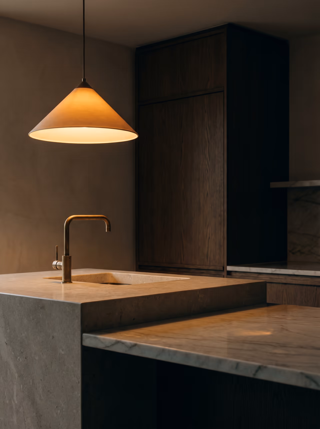 Kitchen at evening — a conical pendant glows over a stone island and marble counter, with a brass gooseneck tap and walnut cabinetry.
