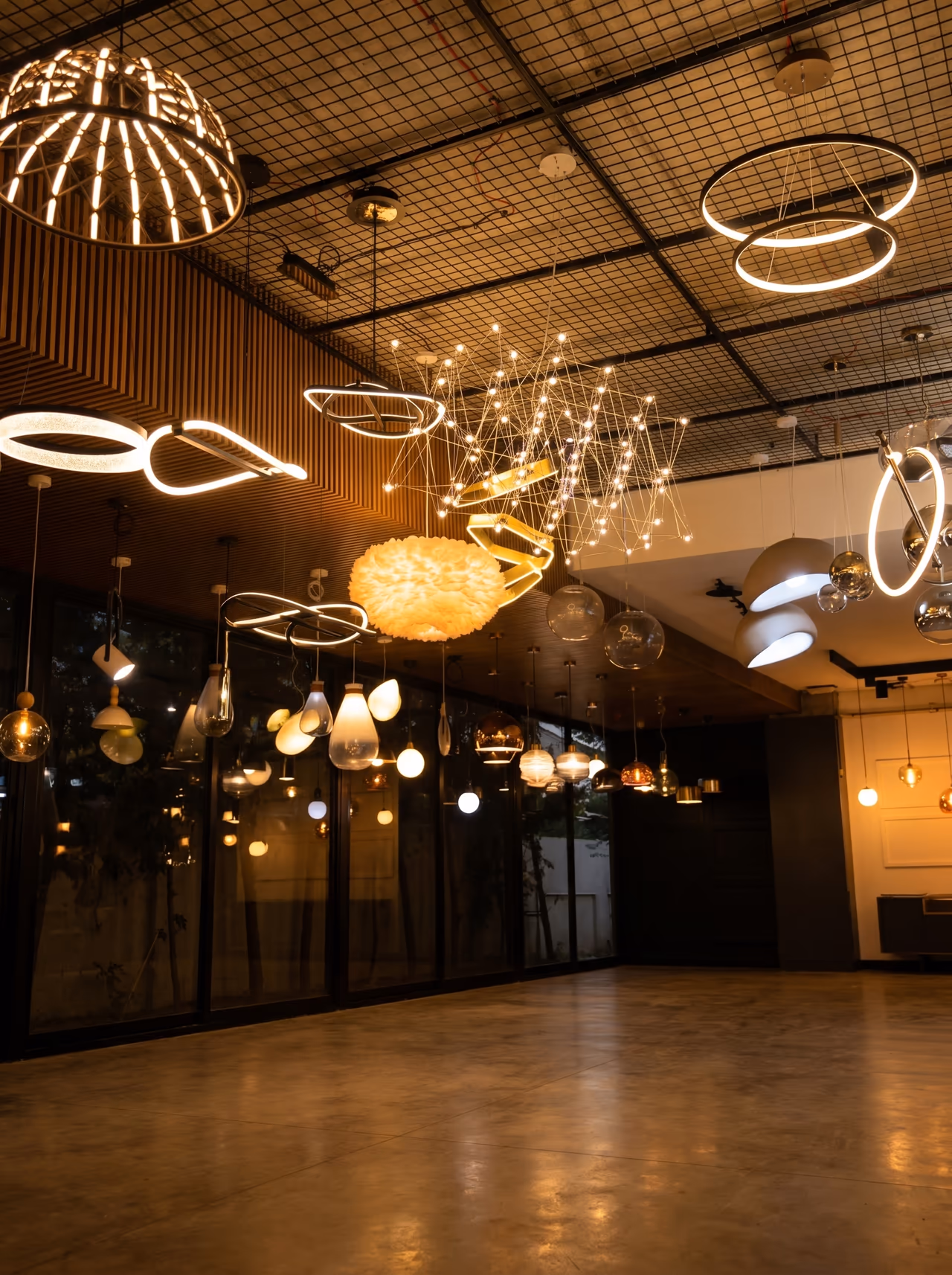 Lighting showroom — glowing decorative pendants, LED ring chandeliers and sculptural fixtures suspended from a grid ceiling.