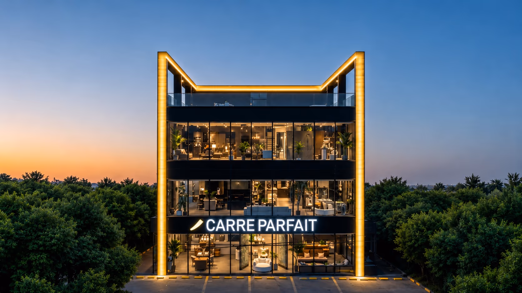 Carré Parfait storefront at dusk, head-on — a glass-fronted building lit by a glowing yellow frame, its illuminated wordmark above the entrance, flanked by dense trees against a soft sunset sky.