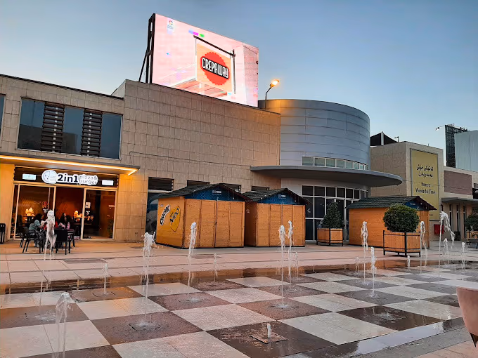 Water-feature plaza at Magma Square at dusk — fountains rising from checkerboard porcelain paving in front of the restaurant row.