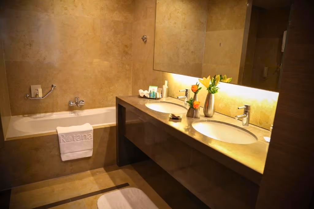 Guest bathroom at the Rotana Erbil — travertine-look wall tiles over a bathtub and a double-basin stone vanity with backlit mirror.