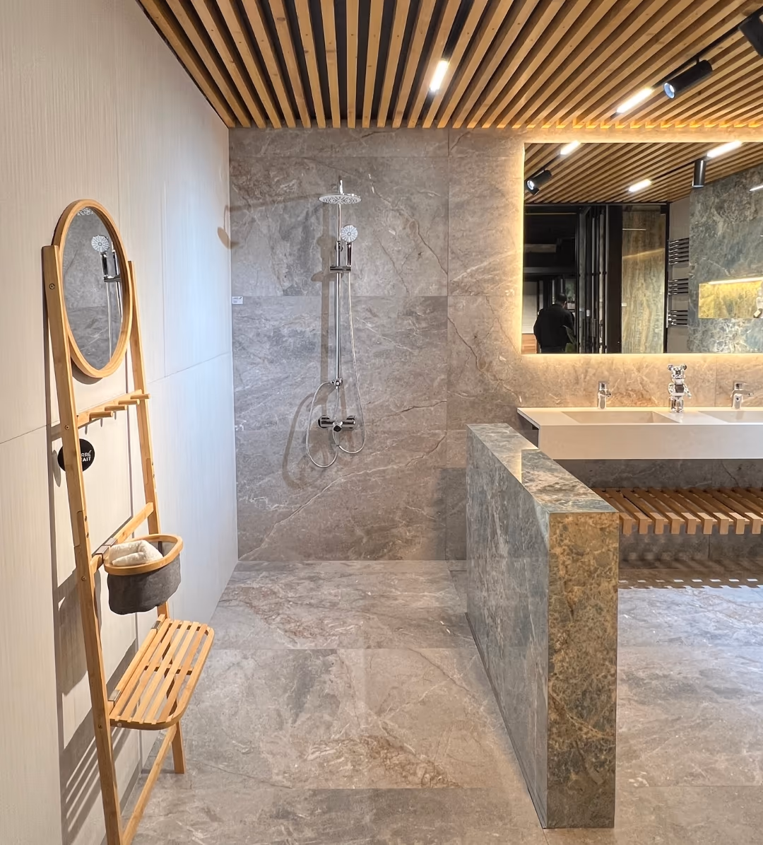 Showroom bathroom display: grey stone-look floor and wall tiles, twin white basins on a green marble-look counter, a bamboo ladder shelf with a round mirror.