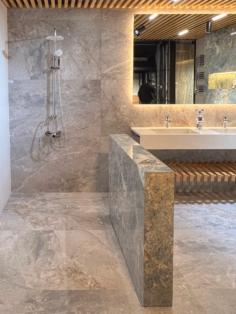 Walk-in shower and white trough vanity in grey marble-pattern large-format tiles, beside a green slab-clad wall and a backlit mirror.