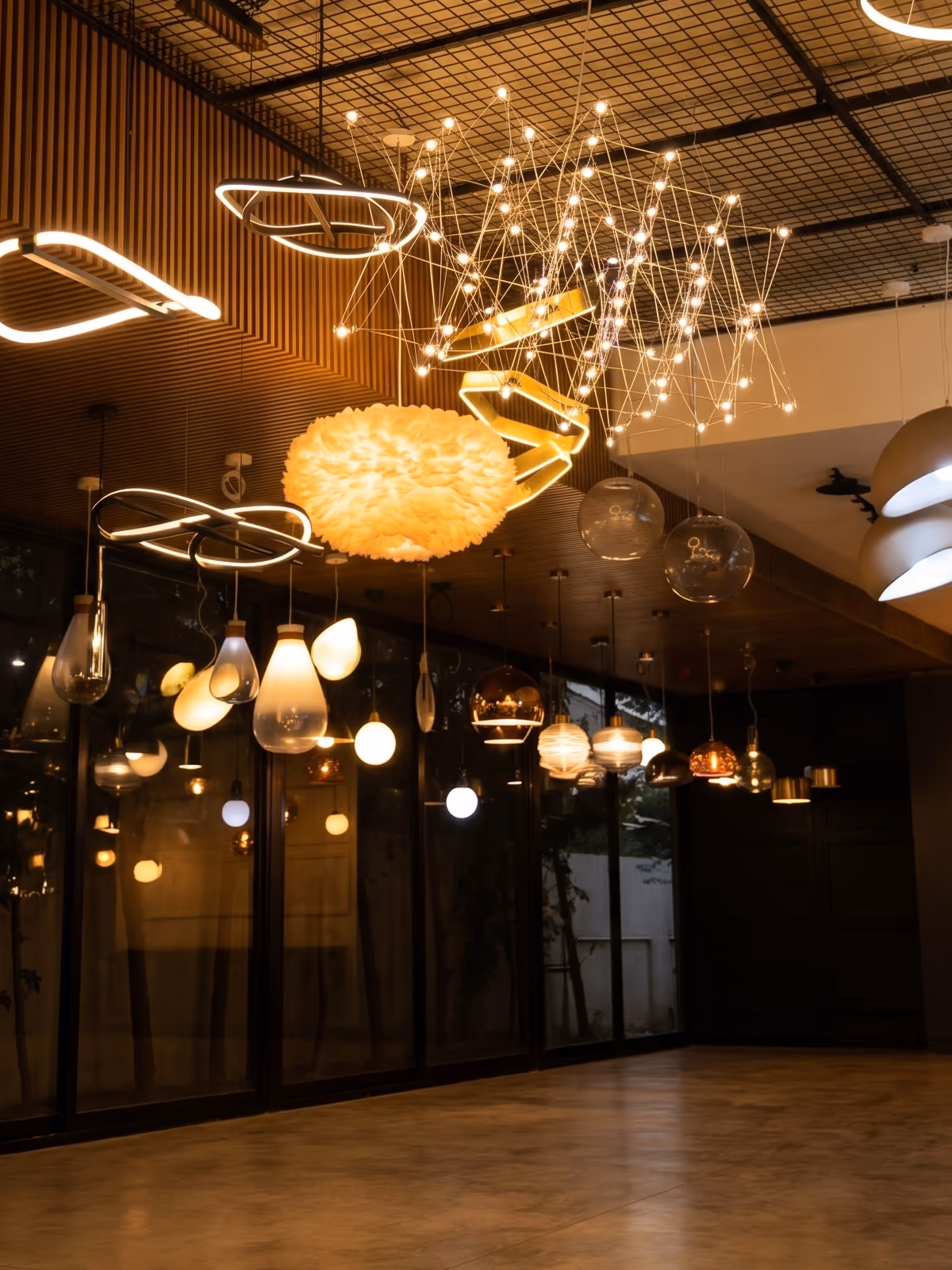 Pendant lights, ring chandeliers and a feather lamp hanging from a black grid ceiling above a polished concrete floor.