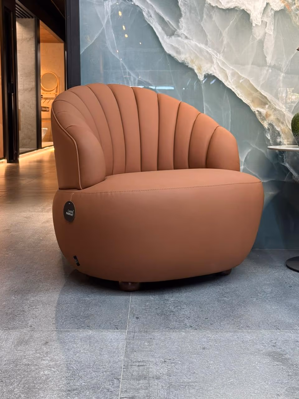 Cognac leather shell-back armchair in front of a teal onyx-pattern slab wall, on grey stone-look large-format floor tiles.